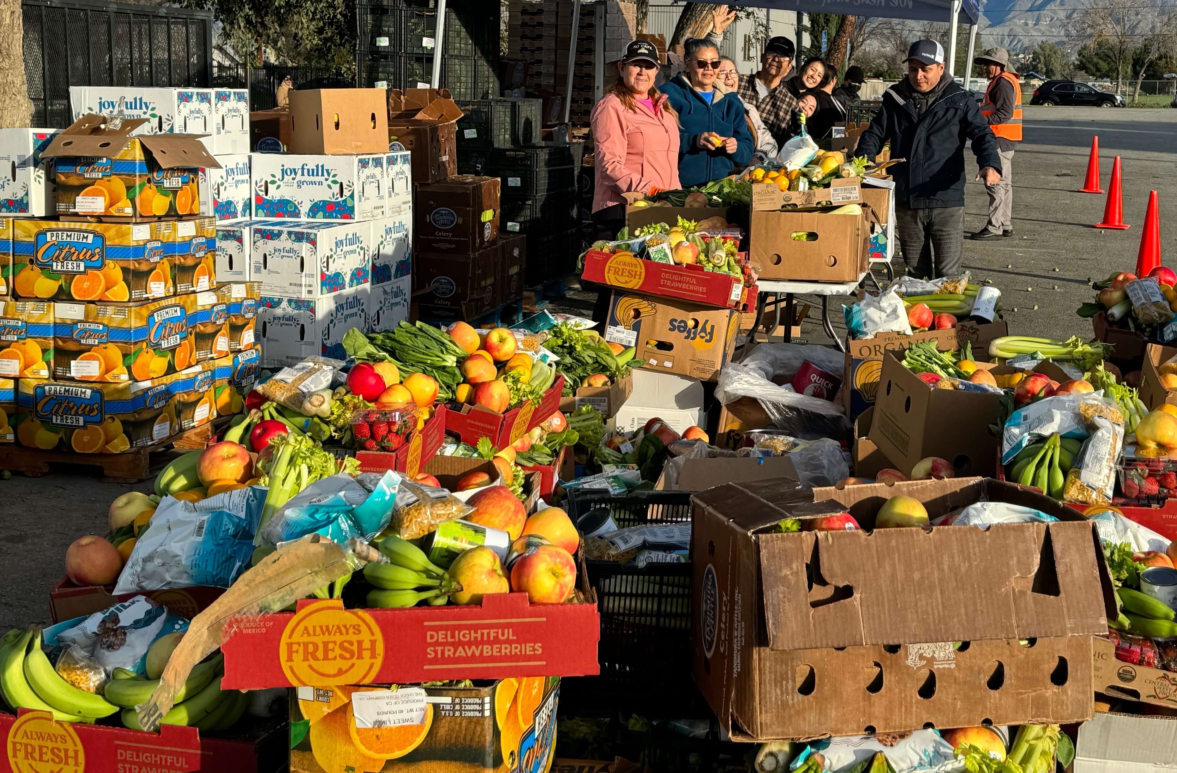 Volunteers stand behind boxes and boxes filled to the brim with apples, bananas, strawberries, celery, other fresh produce at a food distribution.